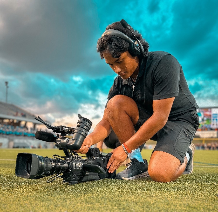 A person with headphones on kneeling on grass with cameraDescription automatically generated
